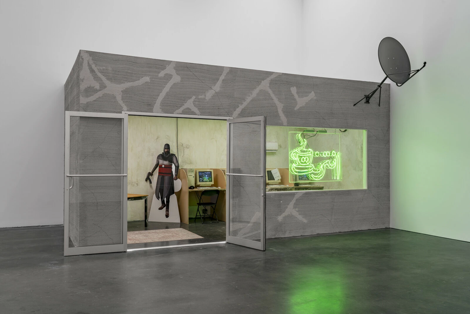 A building in a white gallery space. The outside features repaired cracks, a double door, a neon sign in the window, and a cable dish. Within the building, a cutout and a few computers are visible.