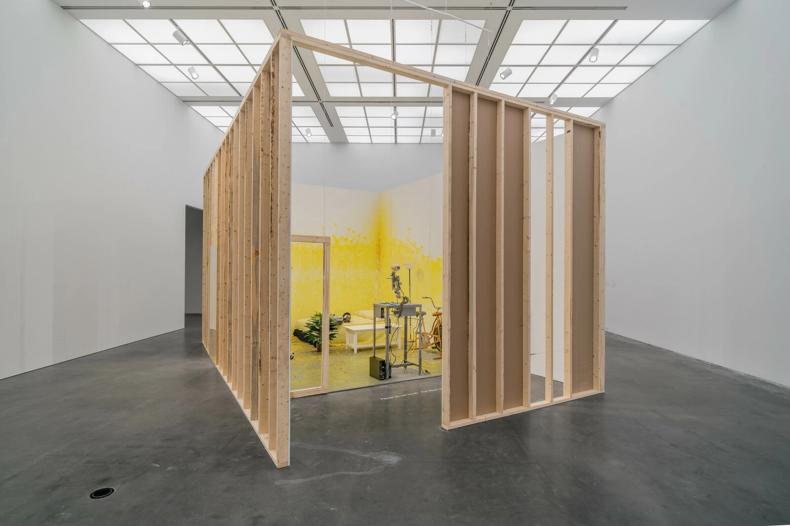 The wooden frame of a room in a large white gallery space. Parts of the room are spraypainted yellow.