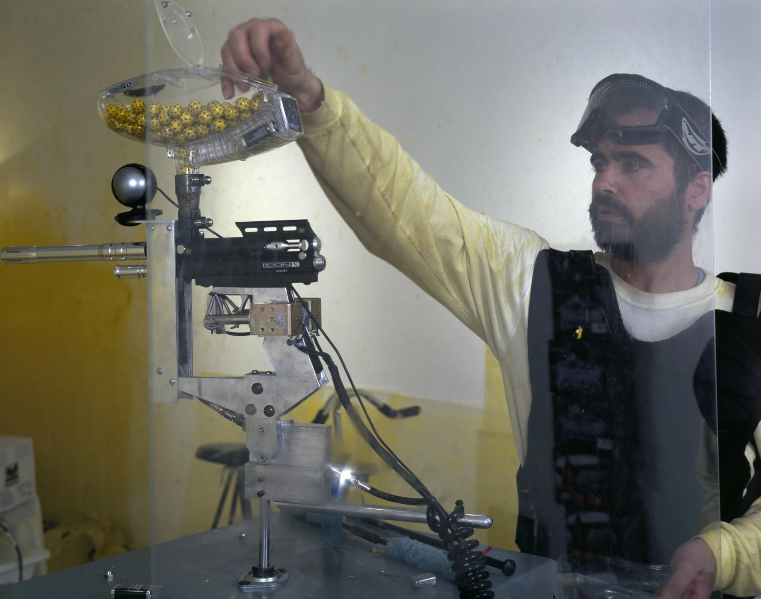 A man with ski goggles and body armor fills an automatic paint gun machine with yellow paint balls.