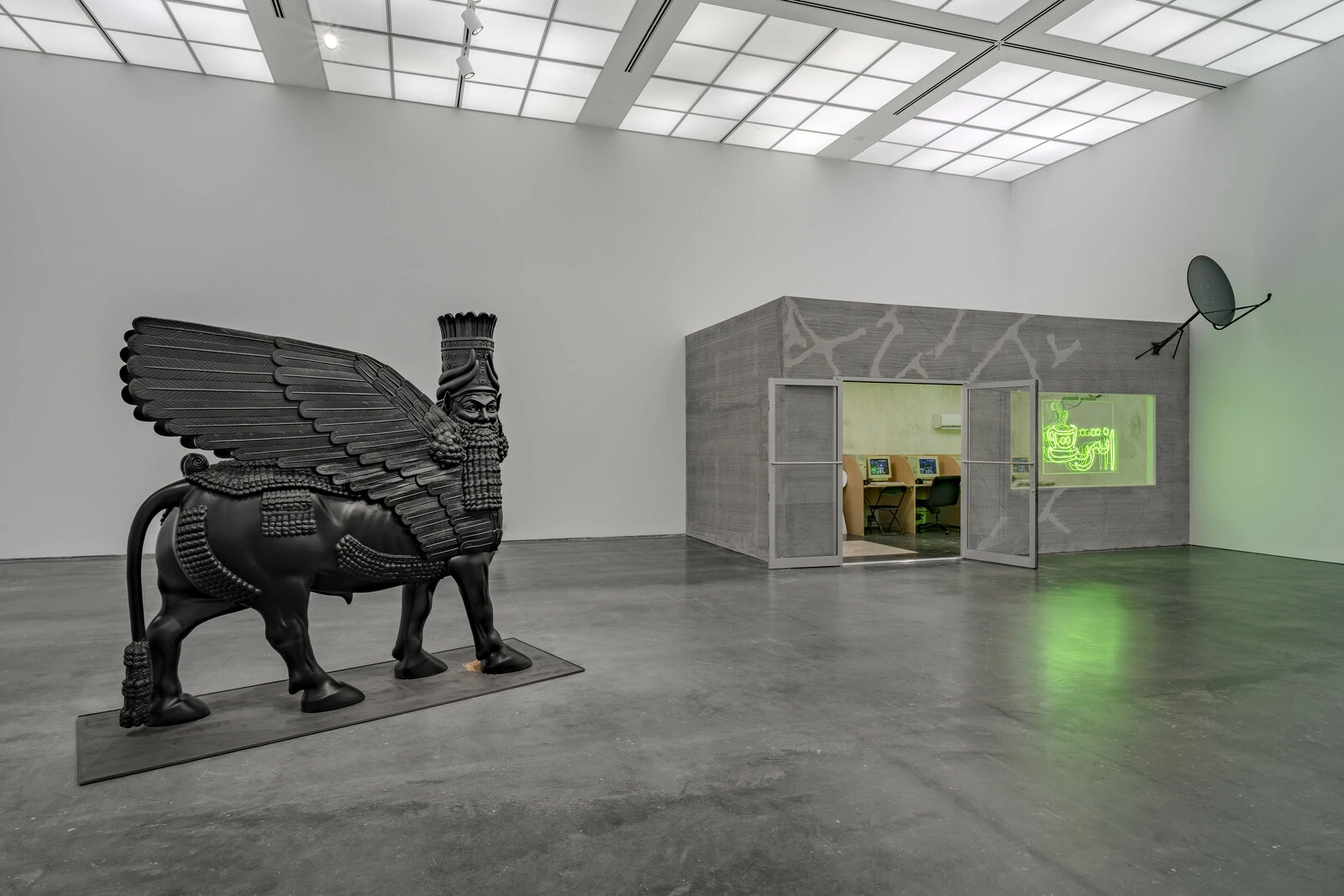 A white gallery space with a large black sculpture in the middle and a room featuring a neon sign appearing in a cutout window and a cable dish in the back corner of the gallery