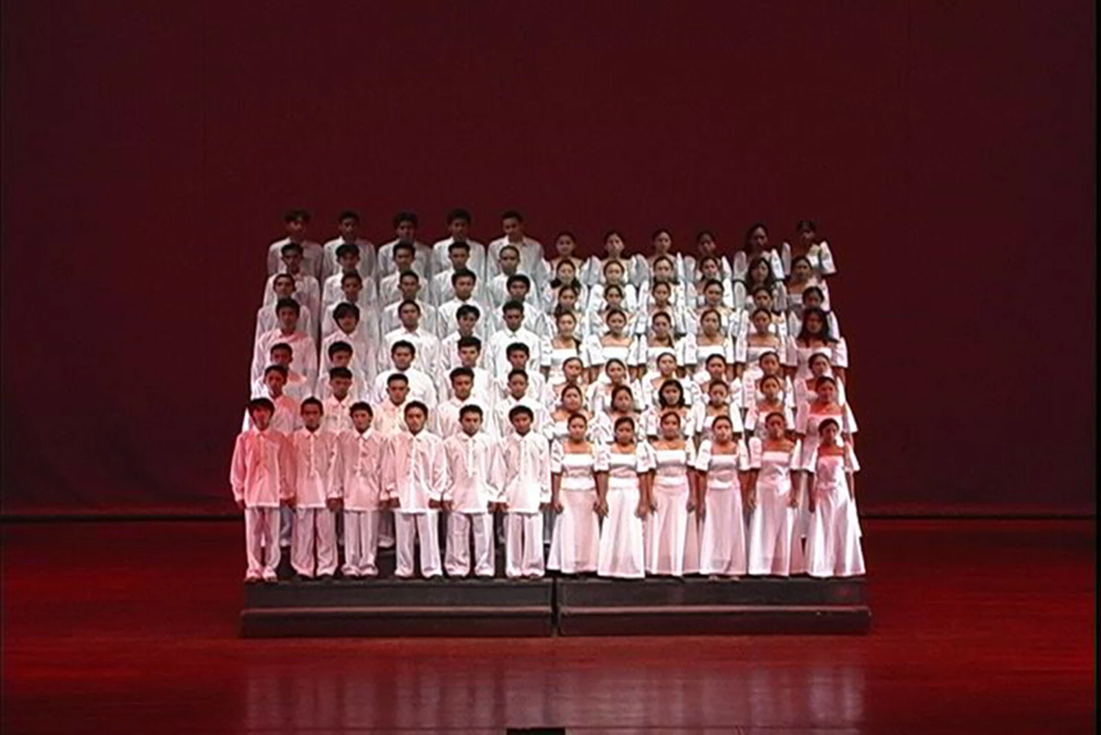 A choir dressed in white on elevated stands on a stage cast in red light