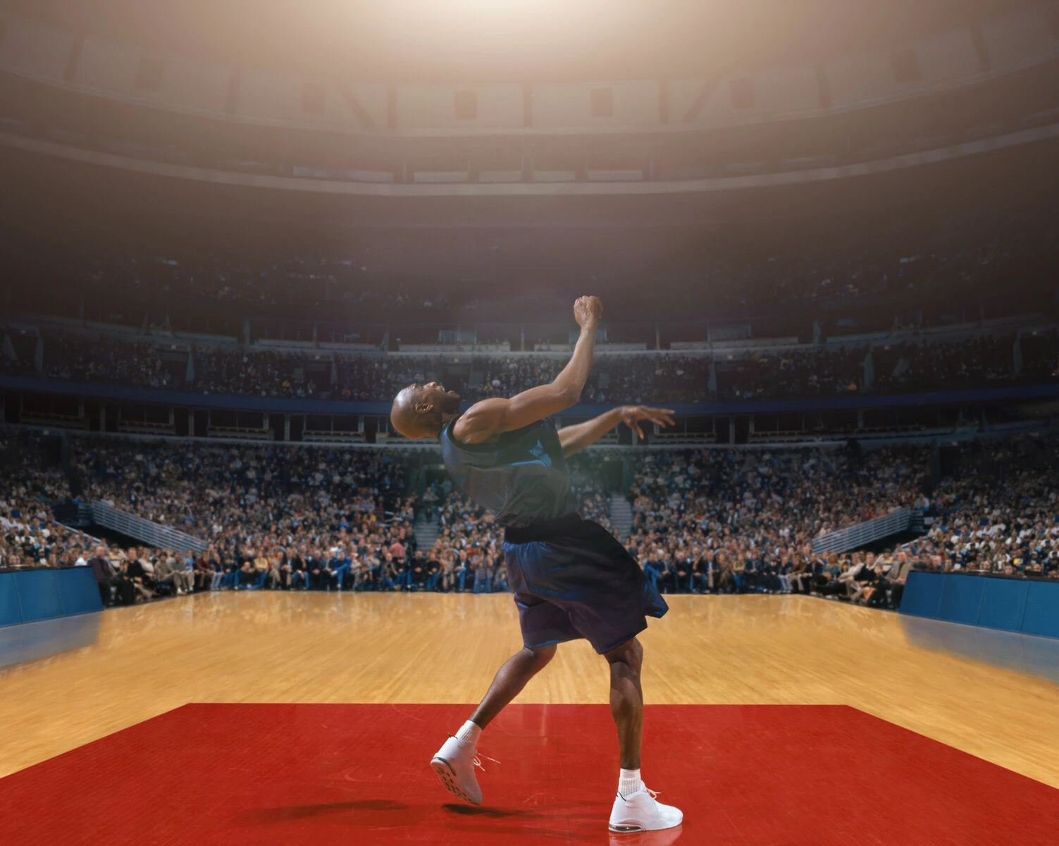 A Black basketball player on a court alone looks like he's about to do a backward flip in front of a crowded stadium.